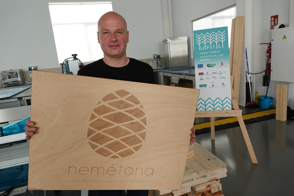 A person is holding a large wooden board with an engraved pinecone design and the word 'nemétona' along with smaller text below it. In the background, there is a sign that reads 'UFIL URBAN FOREST INNOVATION LAB CUENCA' and displays various logos of associated organisations.&nbsp; The setting appears to be a lab with various equipment and tools visible.