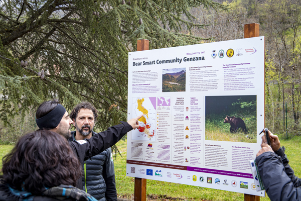 A group of people gathered around an informational sign titled 'Bear Smart Community Genzana.' The sign provides details about the Bear Smart Community initiative, including information on how to coexist safely with bears, the importance of bear conservation, and specific measures to support bear populations. There is a map highlighting areas relevant to the initiative and a photograph of a bear in its natural habitat. The setting is outdoors in a wooded area, suggesting an educational or awareness activity related to wildlife conservation.