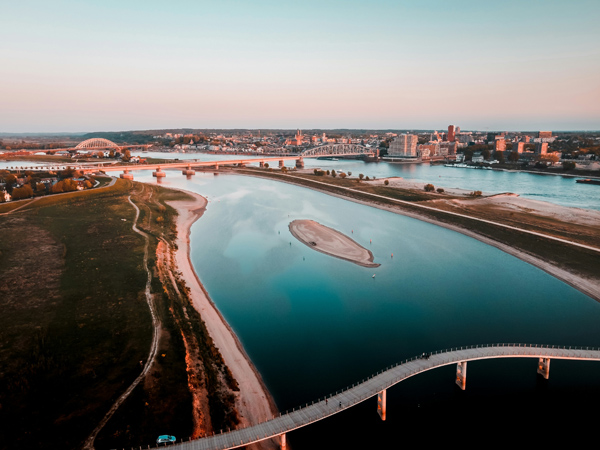Aerial view of a river with a small island in the center, surrounded by green fields and a cityscape in the background. A curved pedestrian bridge crosses the river in the foreground, leading towards the city. The sky is clear with a soft gradient from blue to pink, indicating either sunrise or sunset. The image captures both natural and urban elements, showcasing an interesting blend of landscapes and architecture.