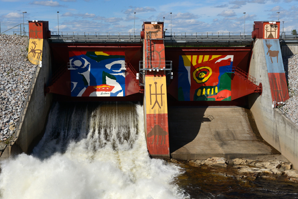 The image shows a dam with water flowing through it. The dam features colorful, abstract murals on its structure.The central mural has a blue and white abstract face with red lips, while the mural on the right side has a yellow and red eye-like design with green and blue elements. The left side of the dam has an image of an animal resembling a deer or elk. There is also some indigenous-style artwork on the central pillar of the dam. The sky above is clear with scattered clouds, and there are rocks piled up along the sides of the dam structure.