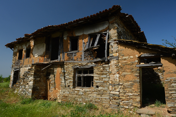 An old, abandoned two-story house with a stone and mud exterior in the Rhodope Mountains, Bulgaria.The structure is dilapidated, with broken windows, missing sections of the roof, and crumbling walls. The surrounding area is overgrown with grass and weeds under a clear blue sky.