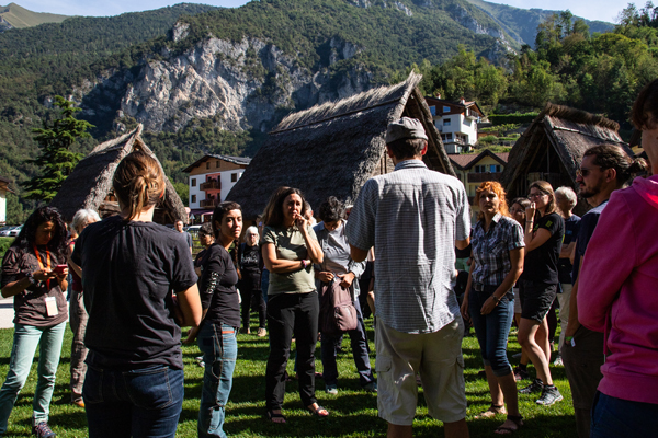 A group of people are gathered outdoors at a community engagement event in Italy focused on coexistence with wolves. The setting is picturesque, with traditional thatched-roof huts and mountainous terrain in the background. The attendees appear to be listening attentively to a speaker who is addressing them from the center of the group.