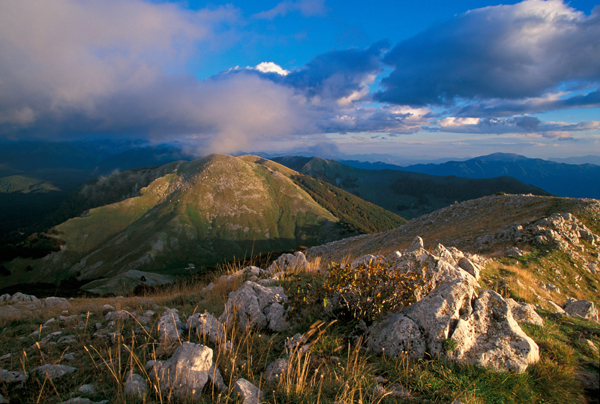 A sunset view of mountain ridges in the Abruzzo National Park, showcasing the characteristic landscape of the Central Apennines in Italy. The image features a rocky foreground with sparse vegetation, rolling green hills, and distant mountain ranges under a partly cloudy sky with dramatic lighting.
