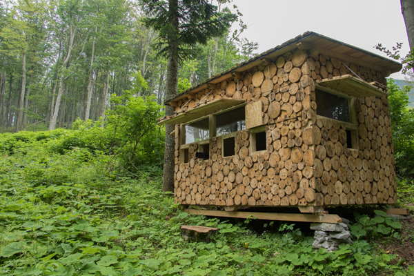 Wildlife watching hide in the Velebit Mountains in Croatia. The photo shows a small wooden cabin in a forested area. The cabin is constructed from stacked logs, with the circular ends of the logs facing outward, creating a rustic and natural appearance. The structure has several windows and appears to be elevated slightly off the ground on wooden and stone supports. The surrounding area is lush with green foliage and tall trees.
