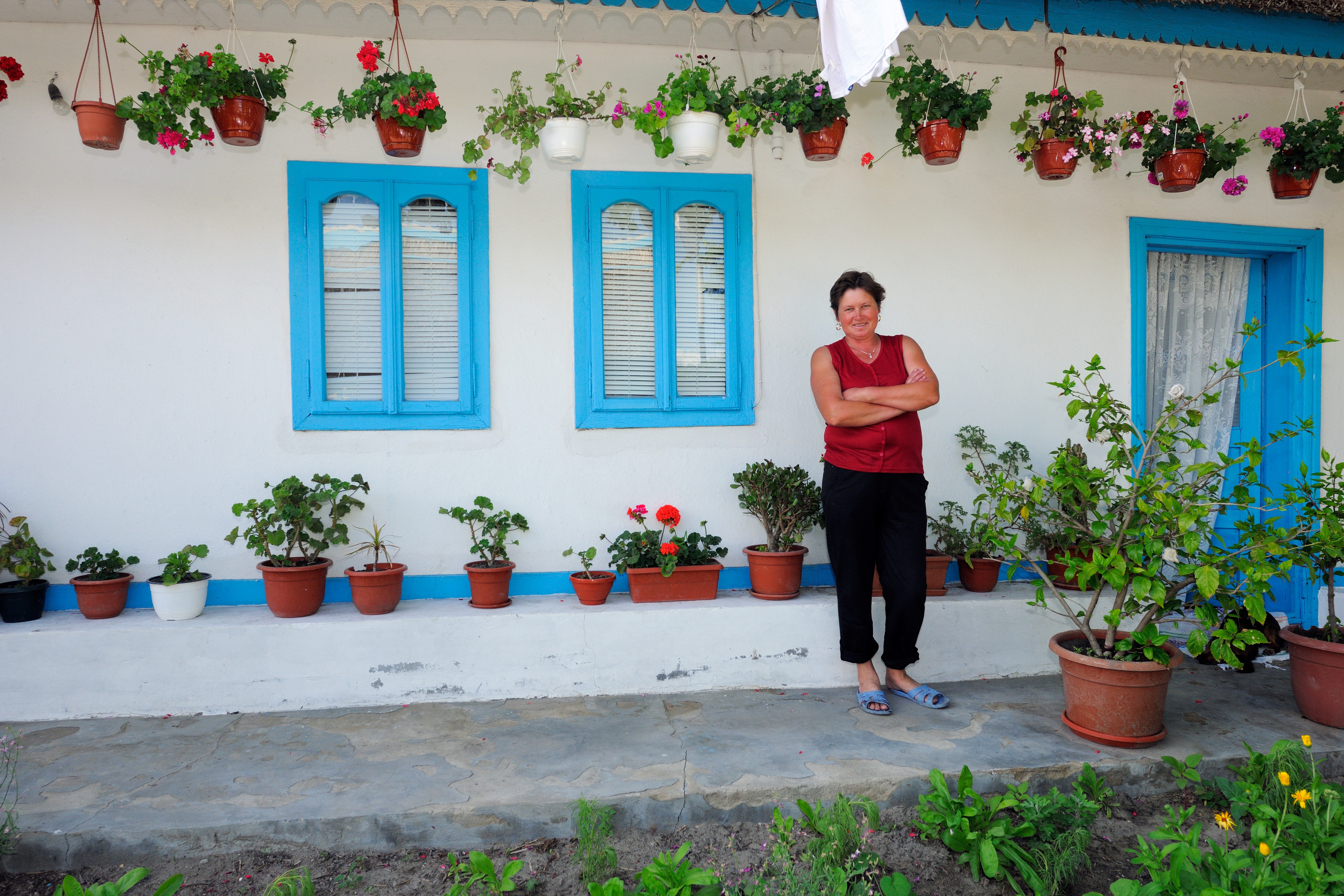 A woman stands with arms crossed in front of a white house with blue window frames and door. The house has several potted plants and flowers hanging from the roof and placed on the ground along the wall.