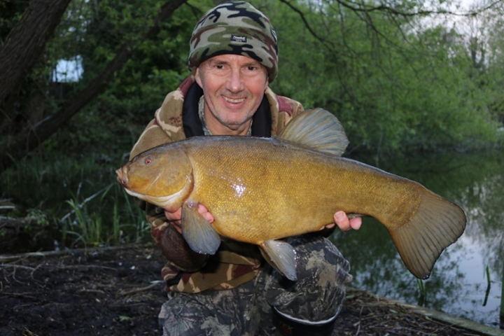 A man standing on a path with a river or lake behind him. The man is holding a large fish with one hand under the fish’s head and the other hand under the start of the tail. The fish is a mottled dark yellow colour with fins and a tail.