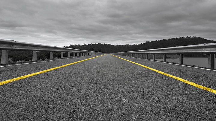 A photograph of a grey tarmac road running vertically in a line from the bottom of the photograph to the horizon. On the left and right hand side of the road are waist-high railings, with a yellow line between the railings and the road.
