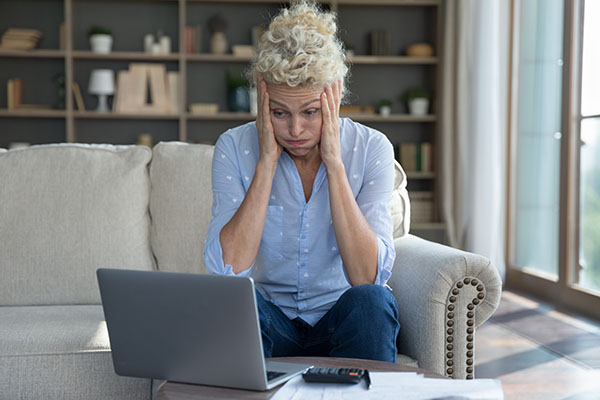 A woman sat on a sofa in a living room with an open laptop on a coffee table in front of here. The woman has her head in her hands and is looking confused.