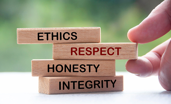 A tower of four wooden rectangular blocks, with one of the following words on each one (from the top down): ethics, respect, honesty, integrity. A hand is holding the second block (from the top) between finger and thumb: it has the word ‘respect’ on it.