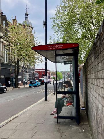 A London bus stop shelter, with a person seated at it. Trees over the shelter and on the other side of the road provide shade