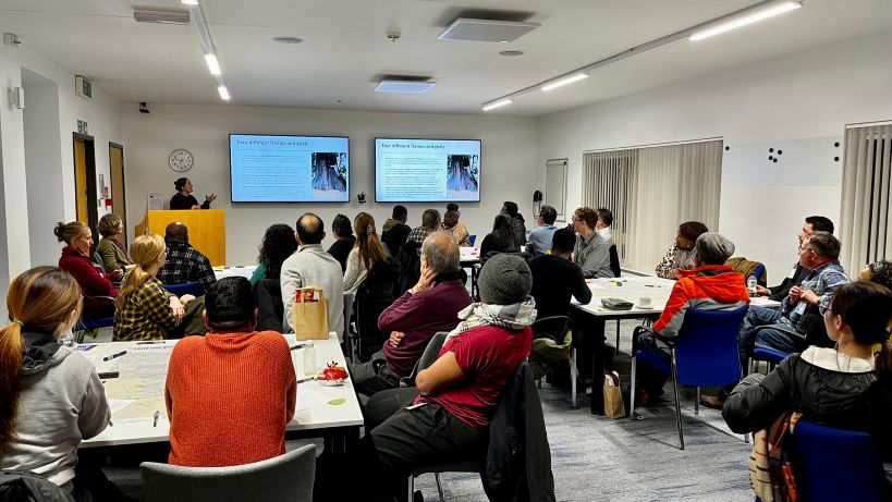 Room with four tables, around 30 people, various genders and ethnicities, looking at a big screen powerpoint presentation