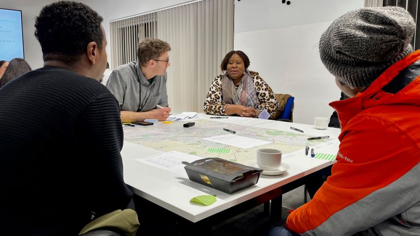 A table in conversation, five people. The table has a map, coffee cups, food, pens, notepads and an audio recording device