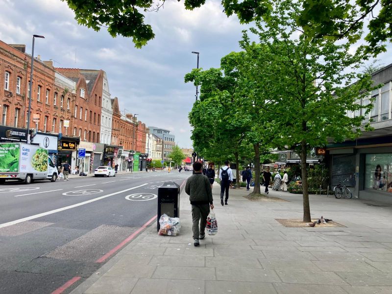 A high street: brick-built shops on the left, post-war arcade on the right. Four traffic lanes & a row of young street trees