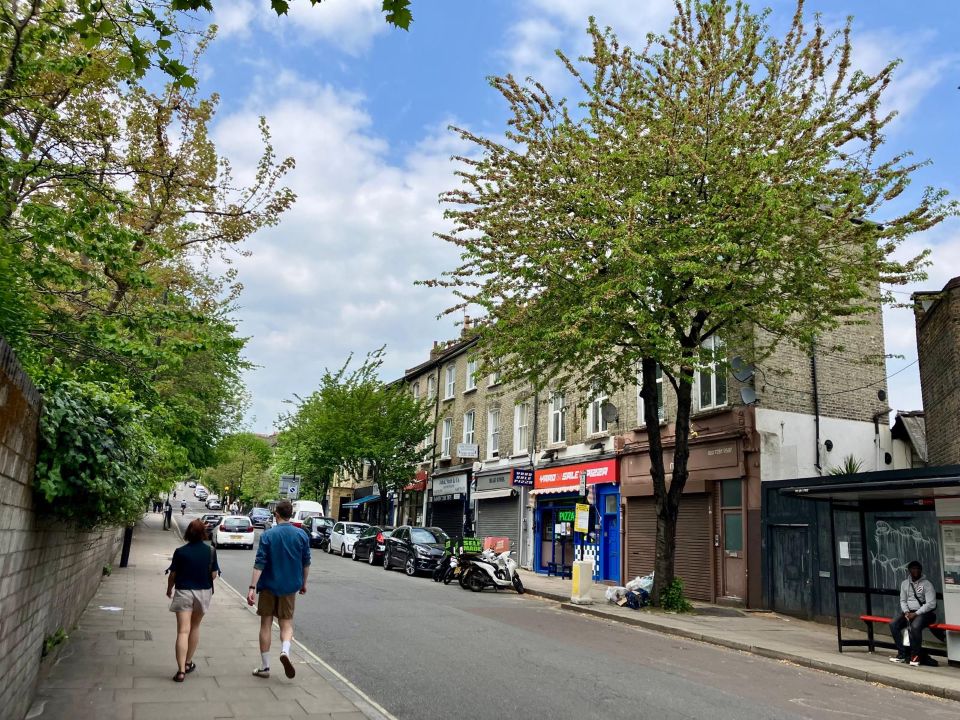 Two people walk down a pavement, a tree is one the opposite side of the road, next to shops, parked cars and a bus stop