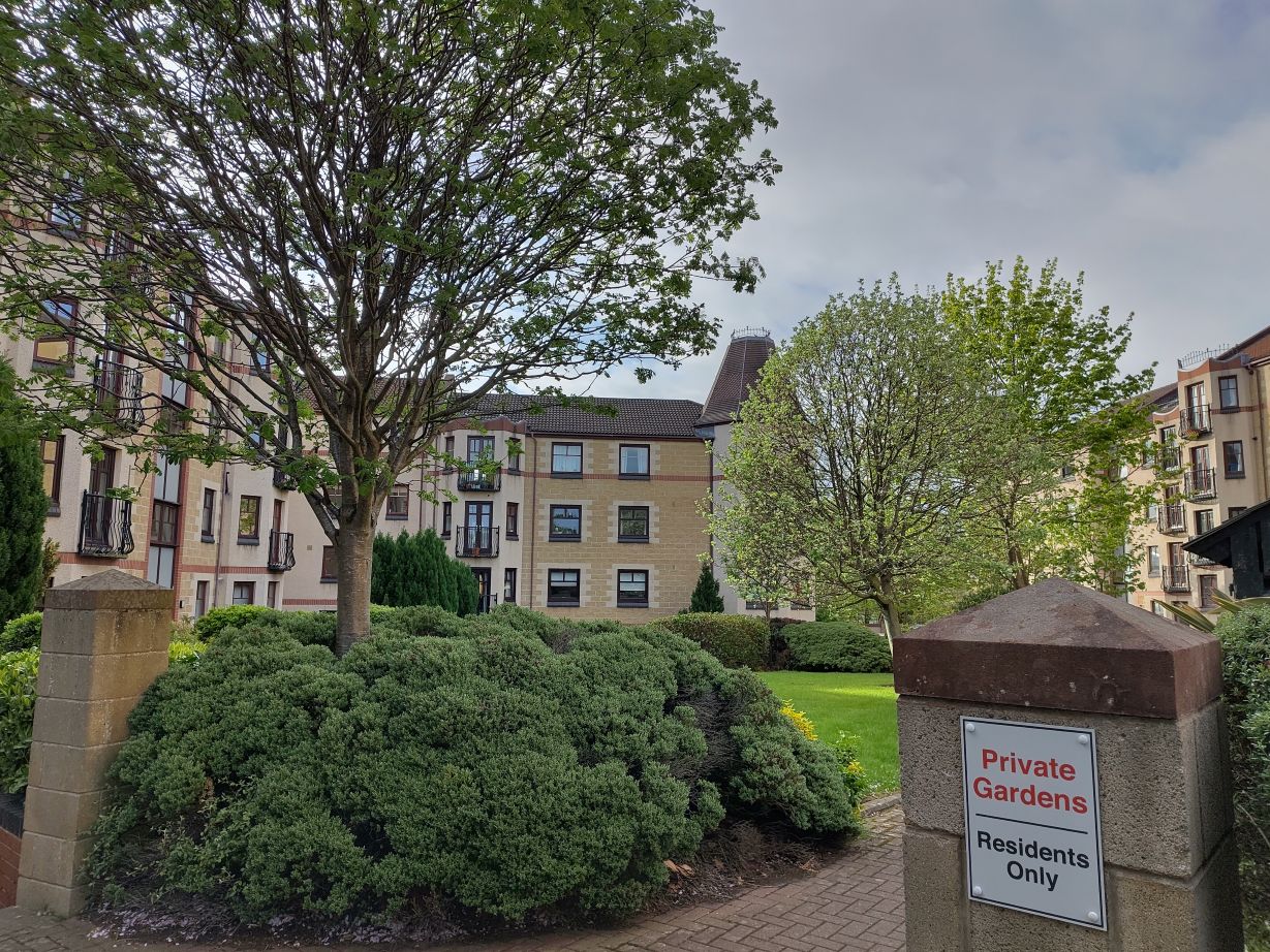 1990s three story flats, with three trees in a communal garden (and bushes). A sign states 'Private Garden | Residents Only'.