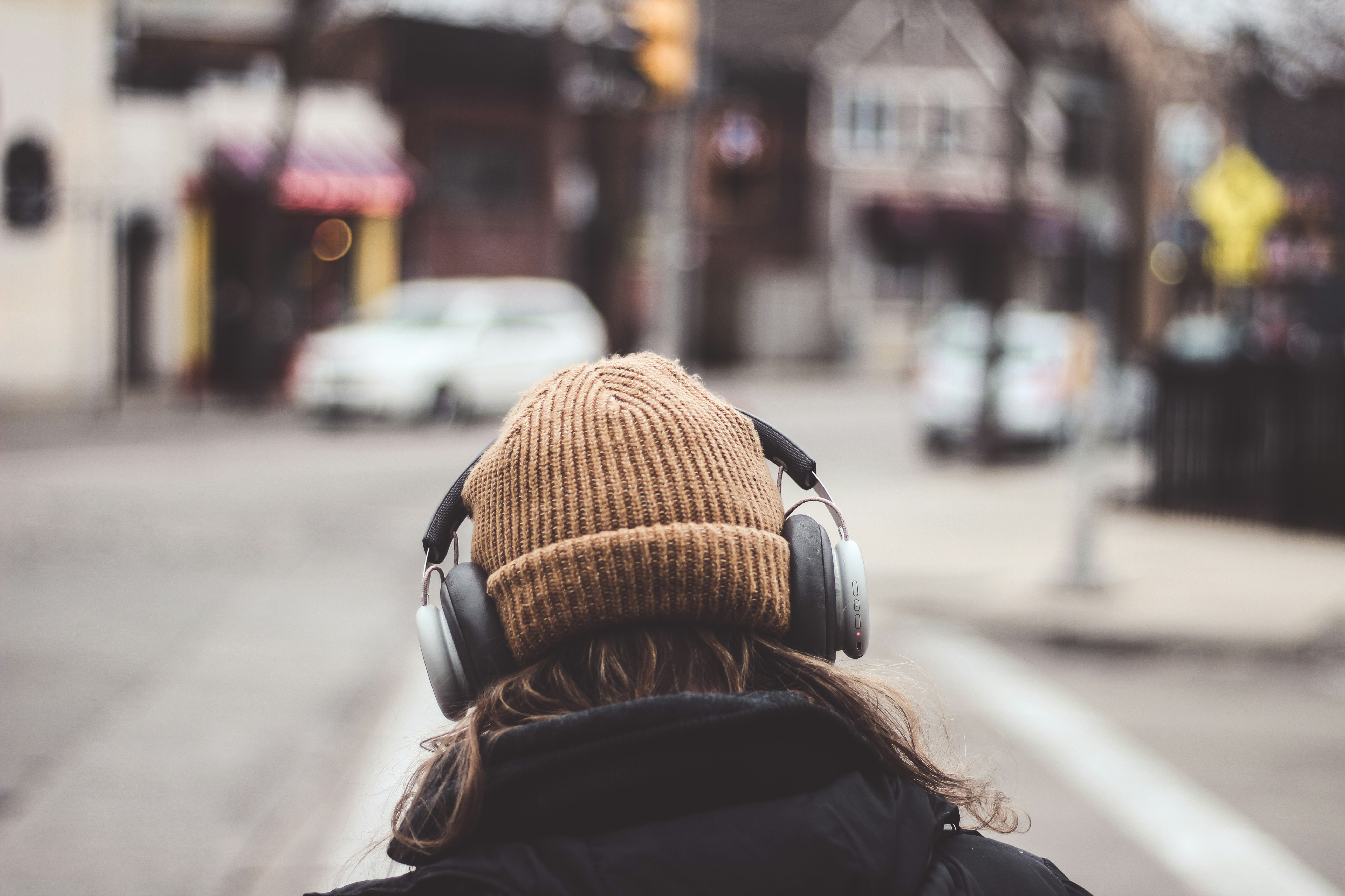A photo taken from behind a person who is walking in the street wearing headphones over a hat and wearing a black coat. 