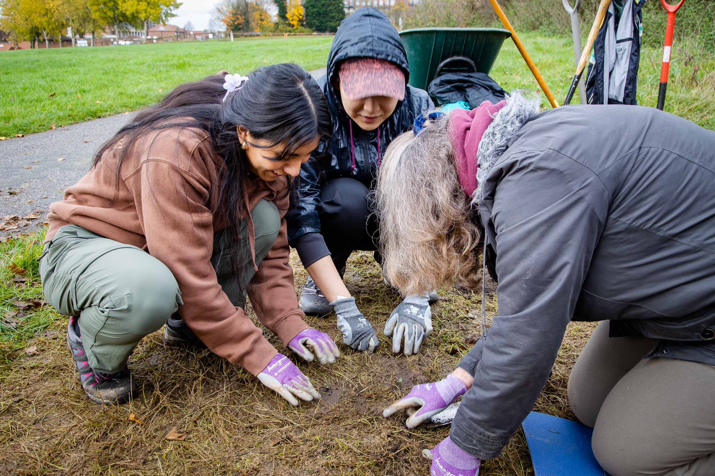Three people are crouching down by a patch of bare soil wearing gardening gloves looking at seeds in the soil.