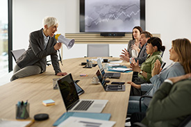 A person standing at the head of a conference table, leaning forward and speaking through a megaphone to a group of seated individuals.