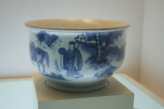A blue-and-white porcelain bowl with a wide mouth and rounded body, painted with scenes of standing figures in robes and stylised trees and rocks, arranged around the exterior surface; the bowl is shown in a museum display setting.