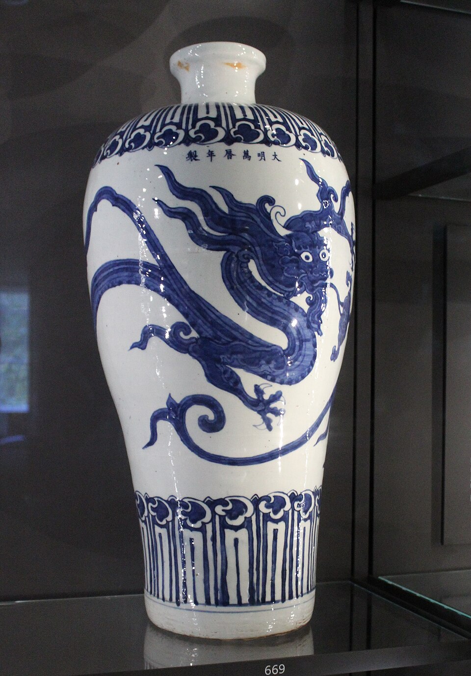 A tall blue-and-white porcelain vase displayed upright, decorated with a large painted dragon winding across the body; the vase has a narrow neck, rounded shoulders, vertical decorative bands near the base, and a white background with cobalt-blue designs.