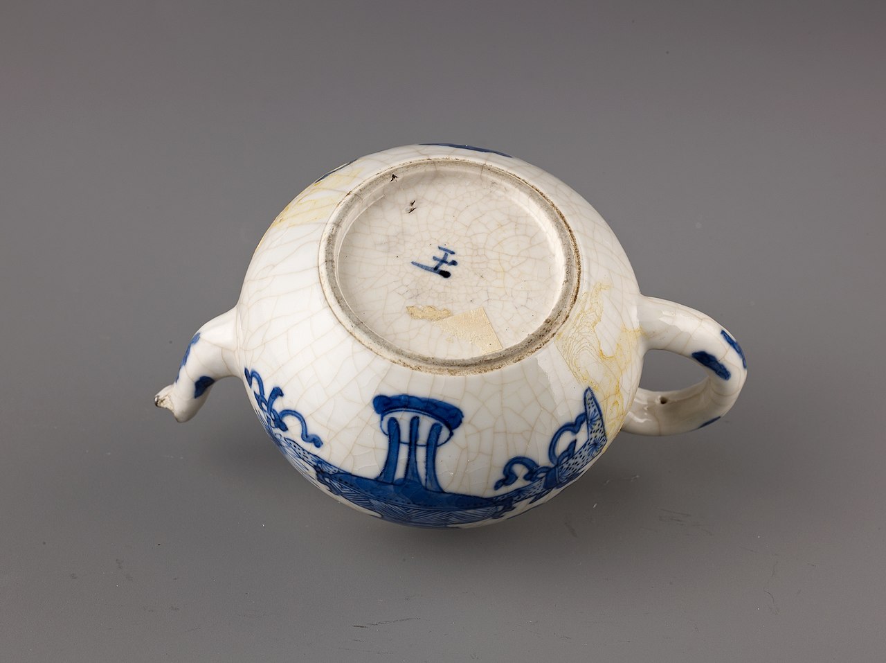 An inverted blue-and-white ceramic wine pot or teapot viewed from the base, showing the foot ring and a blue hand-painted mark at the center; the glaze has visible crackle patterns, with the spout and handle extending on opposite sides.