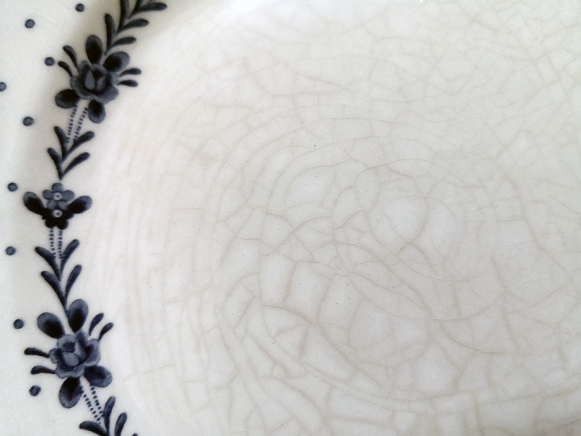 Close-up of a glazed ceramic surface showing fine crackle lines (crazing) across the pale glaze, with a blue painted floral border and small dots along the left edge.