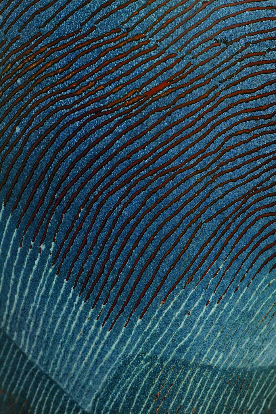 Close-up of a ceramic surface showing layered blue glaze with vertical streaks and dark reddish-brown lines where glaze has flowed downward, creating visible running and pooling effects.