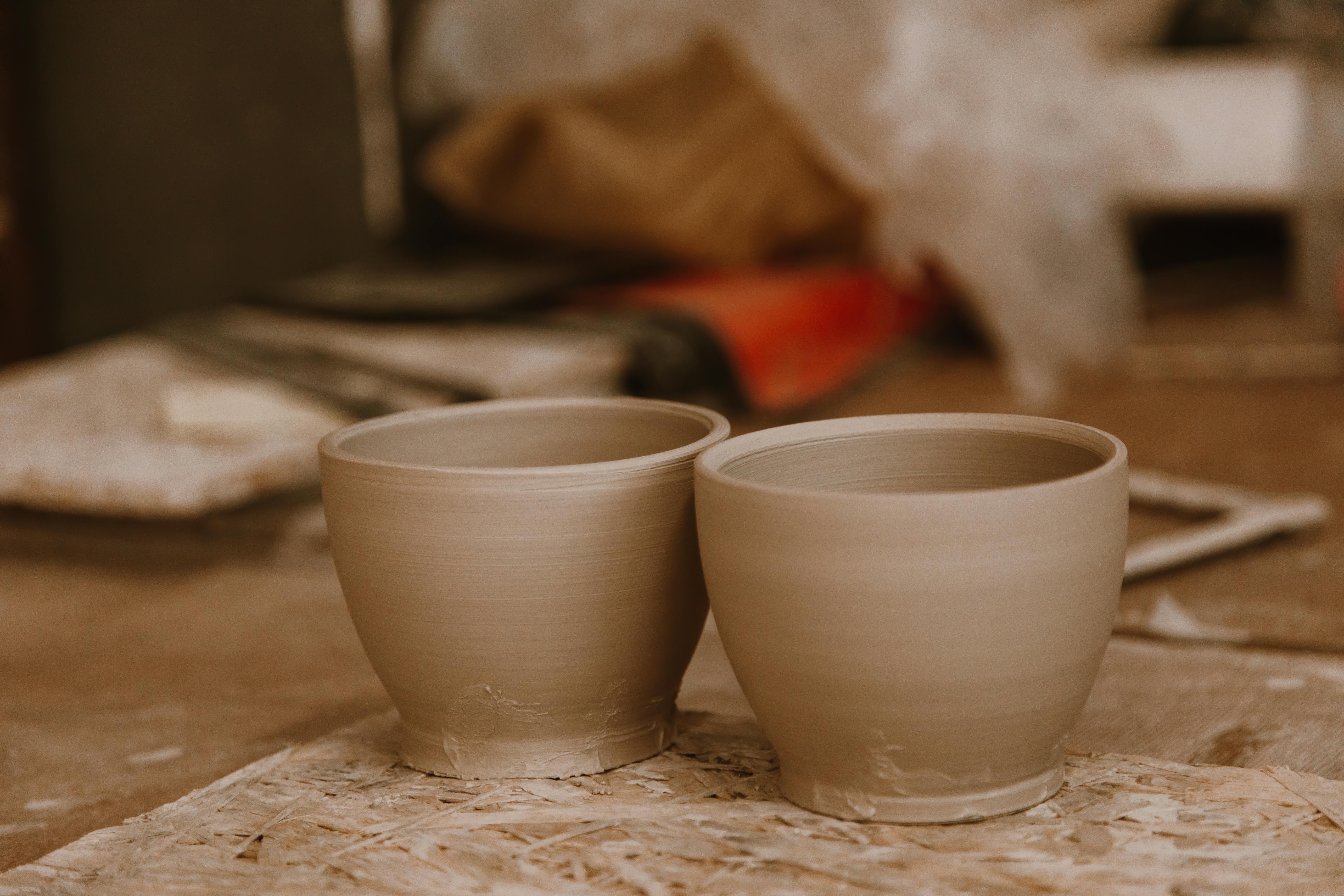 Two unglazed clay cups with smooth, wheel-thrown surfaces sit side by side on a wooden board in a pottery studio, with tools and materials blurred in the background.