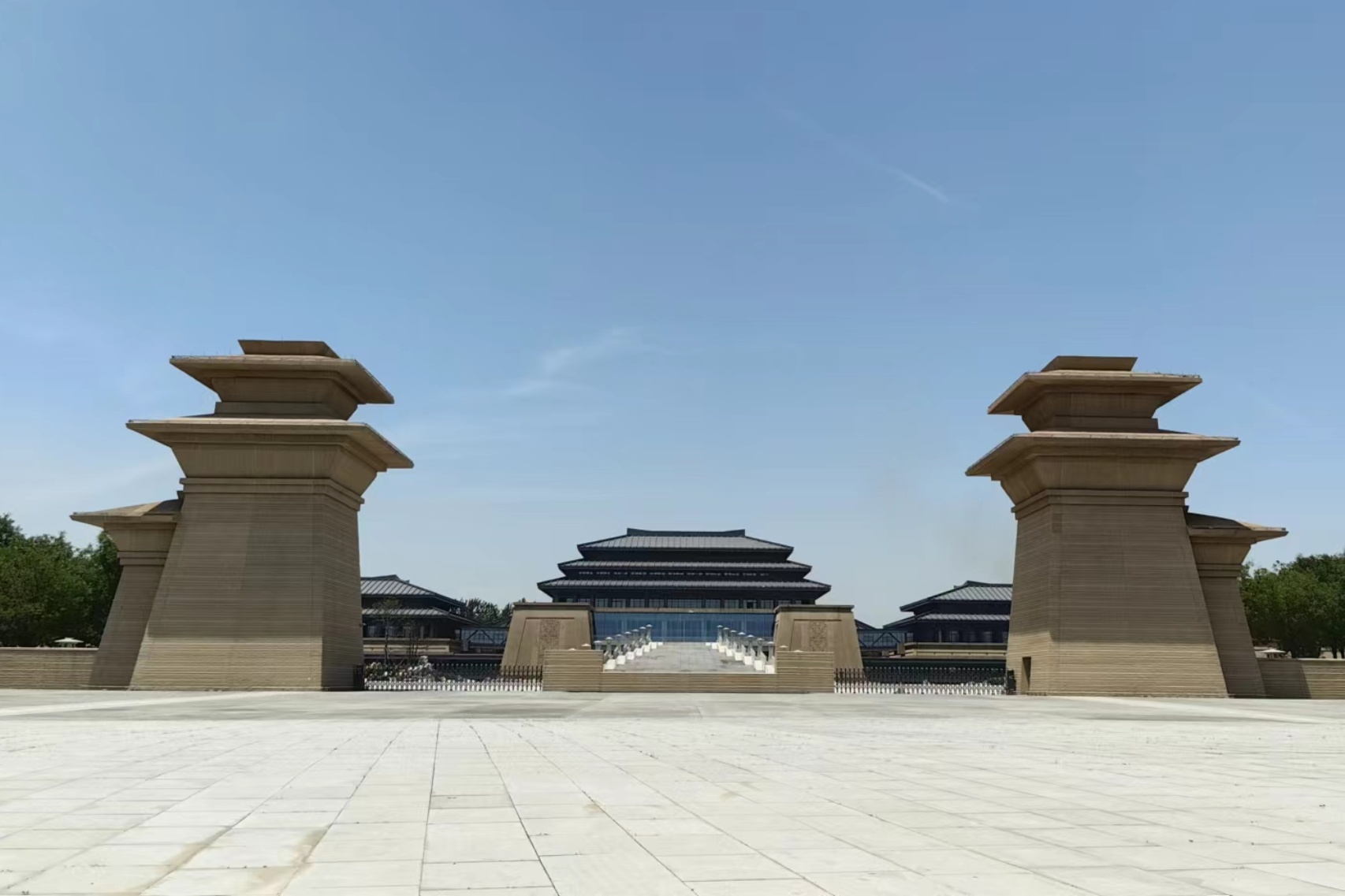 Wide, symmetrical view of the Qin–Han Branch of the Shaanxi History Museum, featuring a large plaza in the foreground, monumental tan stone gate towers on both sides, and a central traditional-style building with layered dark roofs under a clear blue sky.