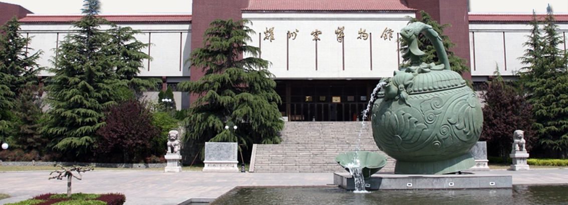 Front view of a museum building with Chinese characters above the entrance, flanked by tall evergreen trees. In the foreground, a large green bronze-style vessel fountain with a sculpted animal handle pours water into a basin within a shallow reflecting pool.