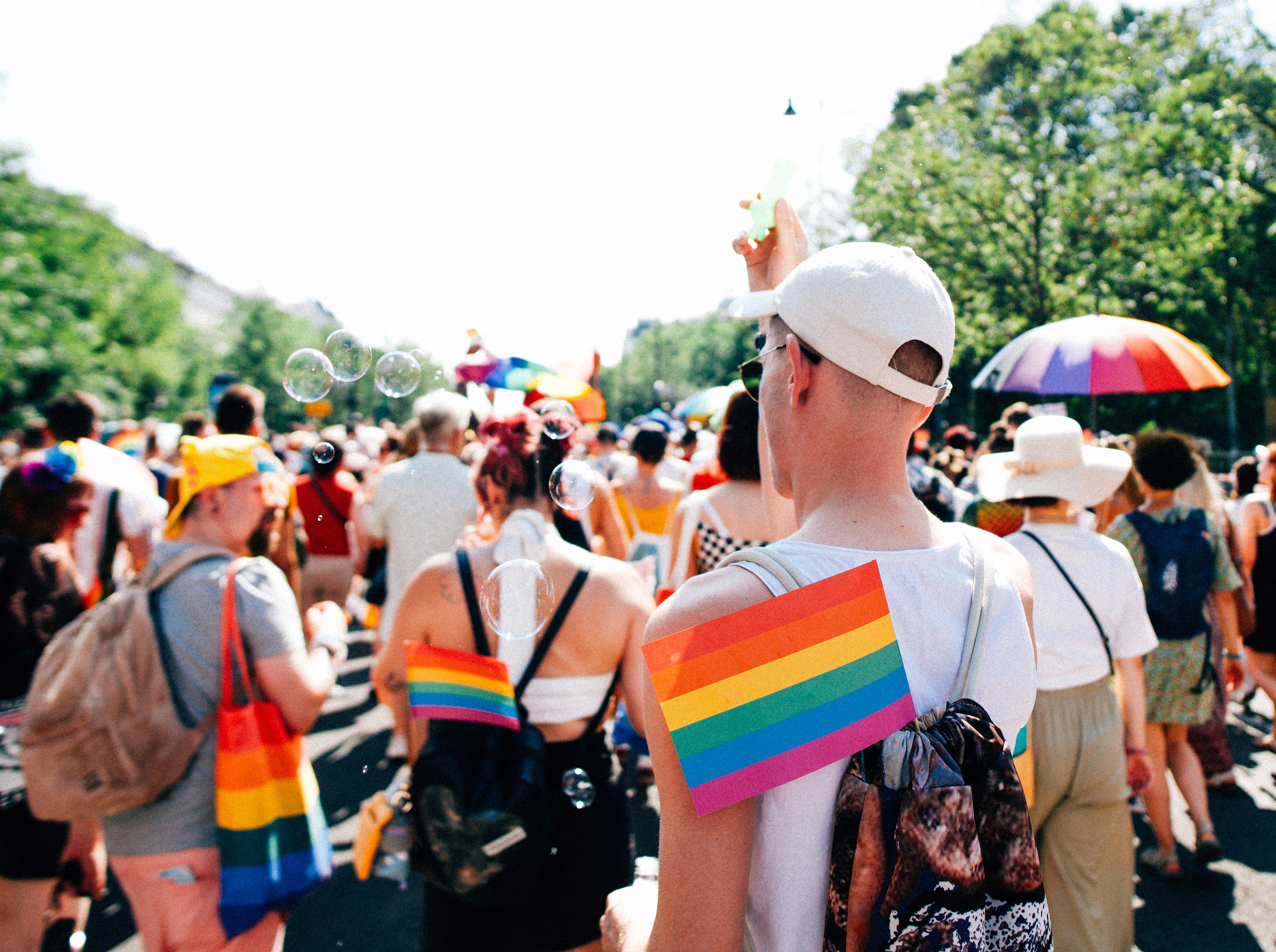 Photo of a pride parade on a sunny summer day. A group of people wearing rainbow-themed accessories is walking down a path in a park.
